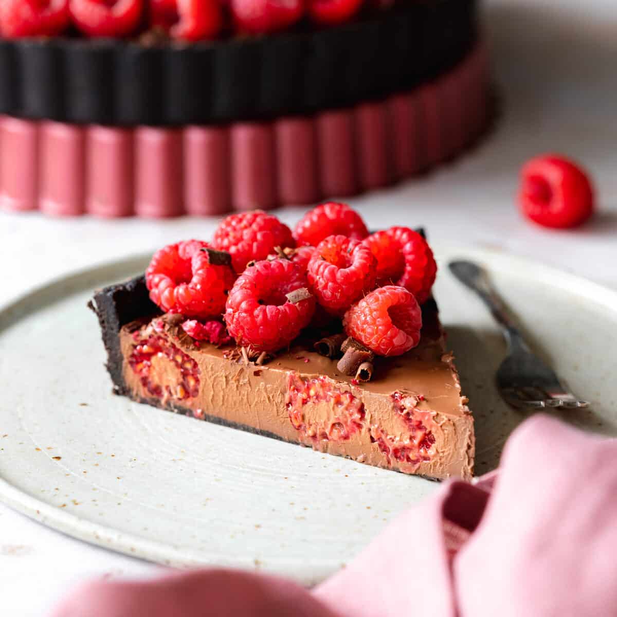 slice of chocolate ganache tart with raspberries on top, and pink linen in the foreground.