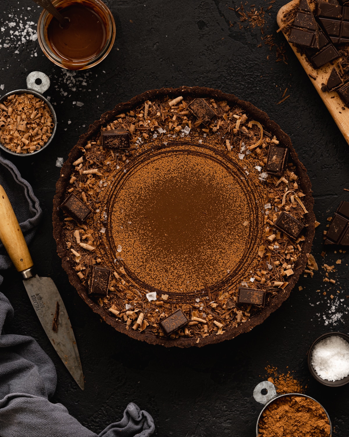flat lay of chocolate tart with cacao powder and chocolate scattered around the scene on a dark background.
